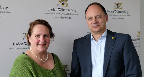 Vor grauem Hintergrund stehen eine Frau in grünem Oberteil, daneben ein Mann mit grauem Jacket. 