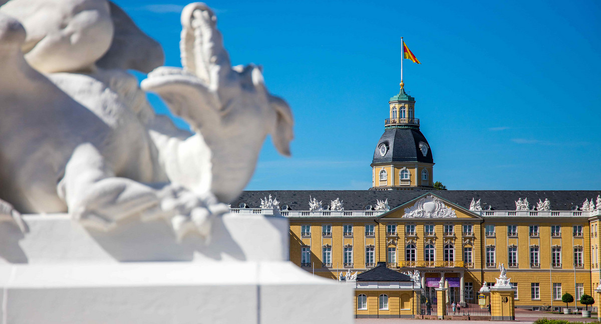 Schloss Karlsruhe mit deutscher Flagge auf dem Dach vor blauem Himmel und unscharfer weißer Skulptur im Vordergrund