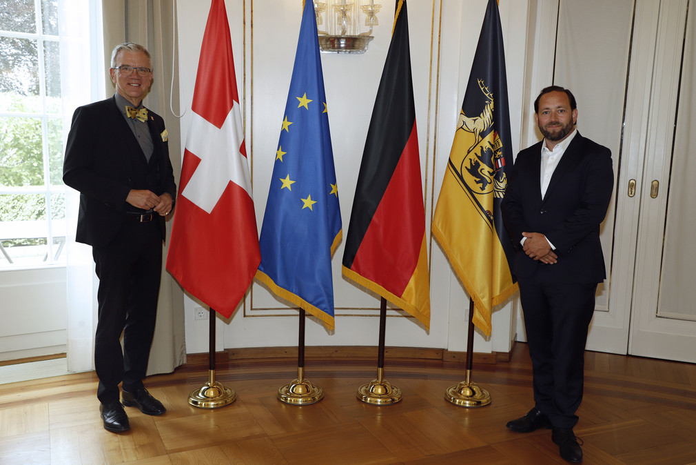 Staatssekretär Florian Hassler (r.) und der Schweizer Botschafter Dr. Paul Seger (l.) stehen neben Fahnen.