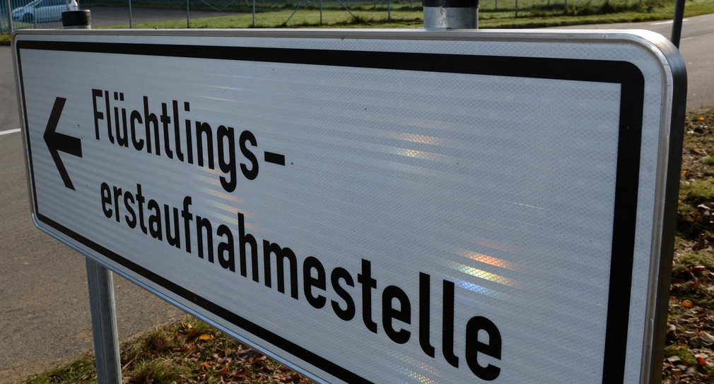 Ein Schild mit der Aufschrift „Flüchtlingserstaufnahmestelle“, im Hintergrund ein Fahrzeug an einer Einfahrt.