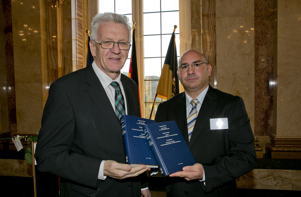 Ministerpräsident Winfried Kretschmann (l.) und Daniel Strauß (r.) halten zwei Bände des Gedenkbuches "Die Sinti und Roma im Konzentrationslager Auschwitz-Birkenau" in der Hand