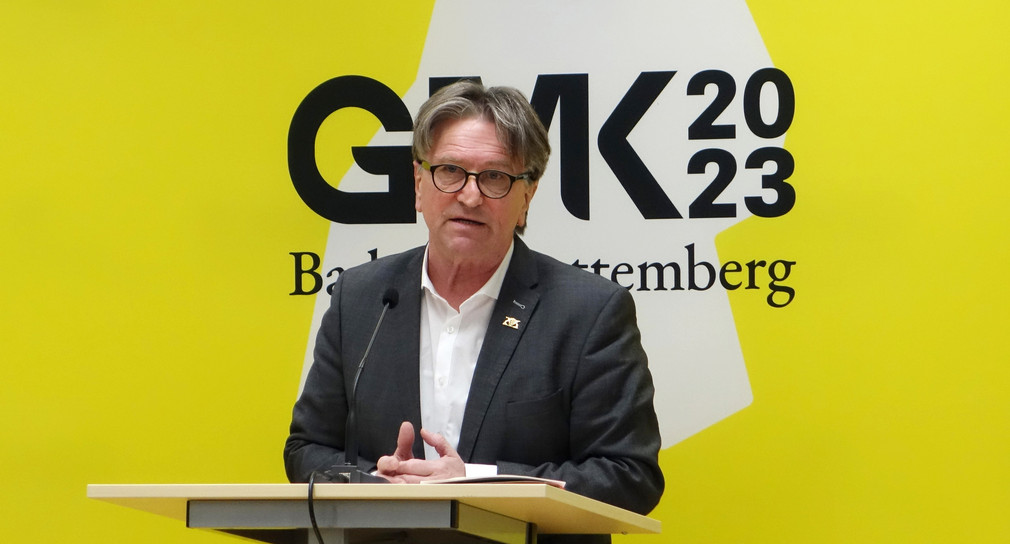 Minister Manne Lucha spricht an Redepult zu Journalisten, im Hintergrund ein großes Banner mit Logo der GMK 2023