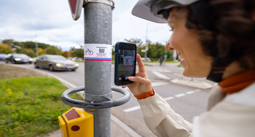 Eine Frau steht an einer Ampel und scant mit ihrem Handy einen QR-Coder von einem Aufkleber der Radkultur. Der QR-Code führt sie direkt zum RadMELDER.