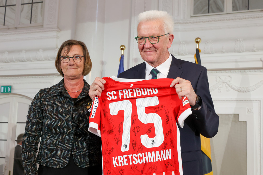Die ehemalige Finanzministerin Edith Sitzmann (links) überreicht Ministerpräsident Winfried Kretschmann (rechts) ein Trikot des SC Freiburg mit der Nummer 75, der Aufschrift "KRETSCHMANN" sowie Unterschriften der gesamten Mannschaft..".