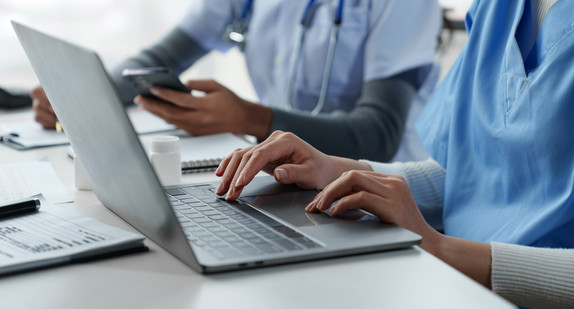 Eine Ärztin und eine Therapeutin sitzen mit einem medizinischen Stethoskop am Tisch und machen mit einem Laptop und einem Mobiltelefon medizinische Notizen.