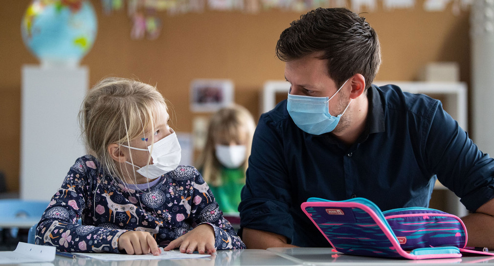 Ein Lehrer mit medizinischer Maske sitzt neben einer jungen Schülerin mit medizinischer Maske.