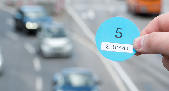 Eine Hand hält einen Designvorschlag des Verkehrsministeriums Baden-Württemberg für die Blaue Plakette in der Innenstadt von Stuttgart vor vorbeifahrenden Autos (Bild: © dpa).