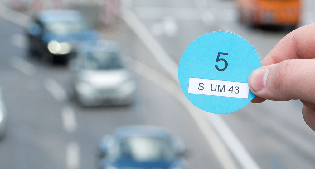 Eine Hand hält einen Designvorschlag des Verkehrsministeriums Baden-Württemberg für die Blaue Plakette in der Innenstadt von Stuttgart vor vorbeifahrenden Autos (Bild: © dpa).