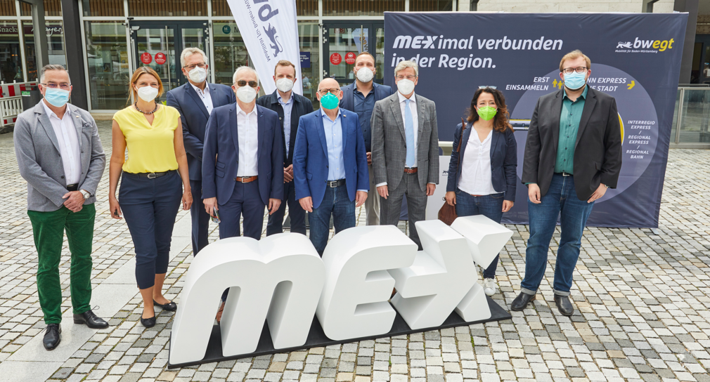 Um große Holzbuchstaben mit dem Titel MEX stehen mehrere Menschen zu einem Gruppenbild zusammen.