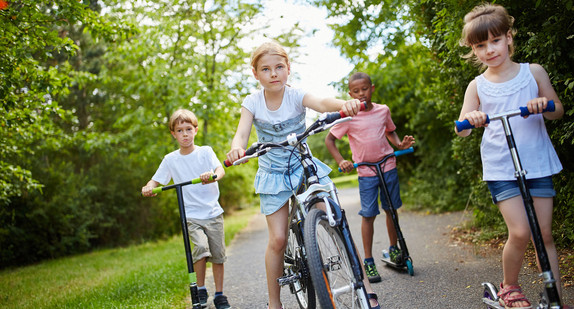 Vier Kinder. Eines auf einem Fahrrad und drei auf Tretrollern auf einem Weg in einem Park.