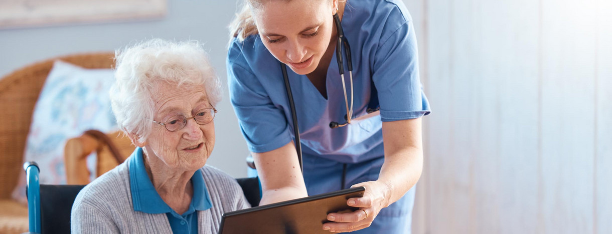 Eine junge Pflegerin zeigt einer älteren Patientin auf dem Tablet medizinische Ergebnisse.