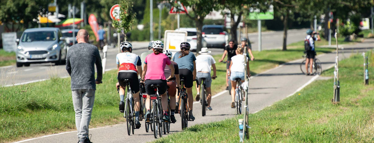 Zahlreiche Menschen sind auf einem Radweg bei Sonnenschein auf ihren Fahrrädern und E-Roller unterwegs.