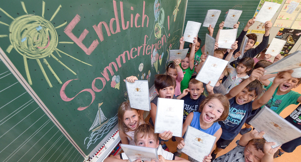 Erstklässler einer Grundschule stehen mit ihren Zeugnissen an der Tafel mit der Aufschrift „Endlich Sommerferien“. (Foto: © dpa)