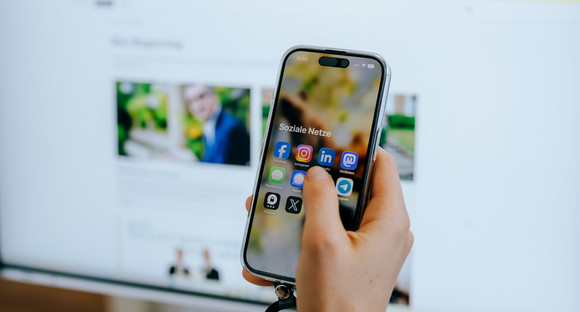 Jemand sitzt vor einem Computerbildschirm und hält ein Smartphone in der Hand auf dem mehrere Social-Media-Apps zu sehen sind.
