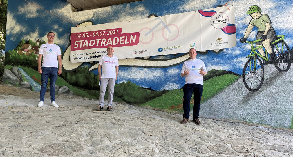 Drei Männer stehen vor einem Graffiti das von einem Plakat verhüllt ist, auf dem für die Aktion Stadtradeln geworben wird.