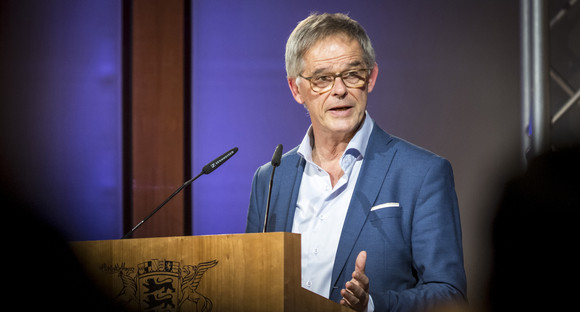 Staatssekretär Rudi Hoogvliet begrüßt die anwesenden Gäste vom Rednerpult aus. Er trägt ein hellblaues Hemd und ein blaues Sakko. Seine Brille ist dunkelbraun meliert. Auf der Vorderseite des Rednerpultes aus Holz sieht man teilweise das Wappen Baden-Württembergs. Der Hintergrund ist in den Farben des Key Visuals beleuchtet: Dunkelblau-Violet.