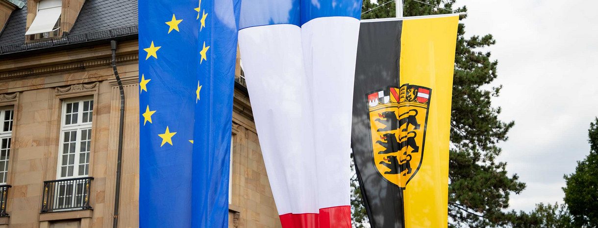 An Fahnemasten hängen die Fahne der EU, die französische Fahne und die baden-württembergische Fahne.