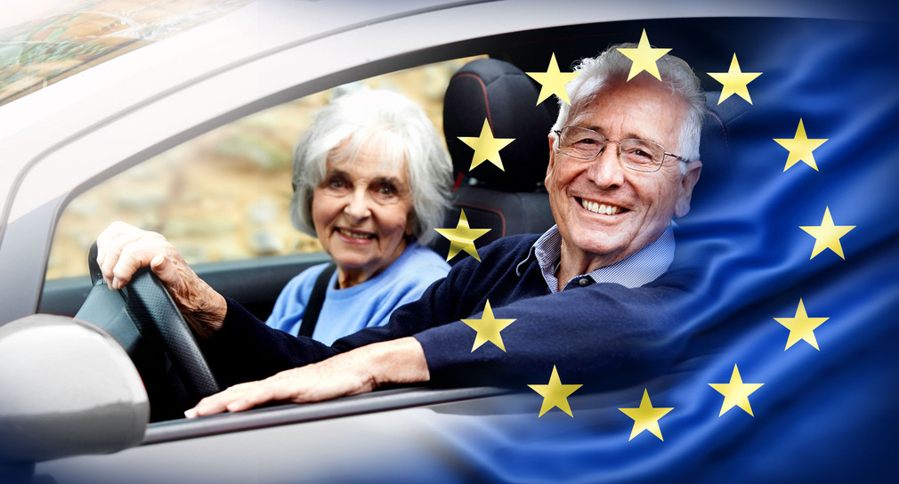 Ein älteres Paar sitzt lachend im Auto. Im Vordergrund ist eine halbtransparente EU Flagge.