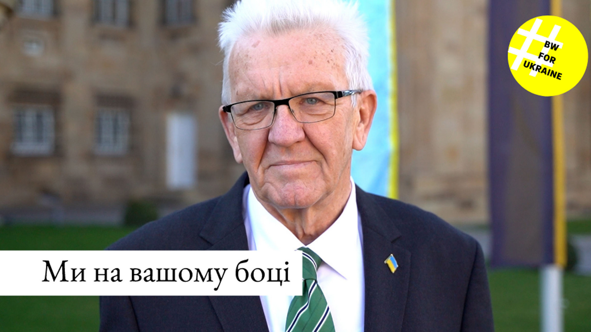 Ministerpräsident Winfried Kretschmann steht bei einer Ansprache zum Jahrestag des russischen Angriffskrieges gegen die Ukraine vor der Villa Reitzenstein in Stuttgart. Im Hintergrund sind die ukrainische und die baden-württembergische Flagge zu sehen.