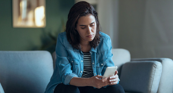 Eine junge Frau sitzt auch einem Sofa und schaut traurig auf ein Smartphone in ihrer Hand.