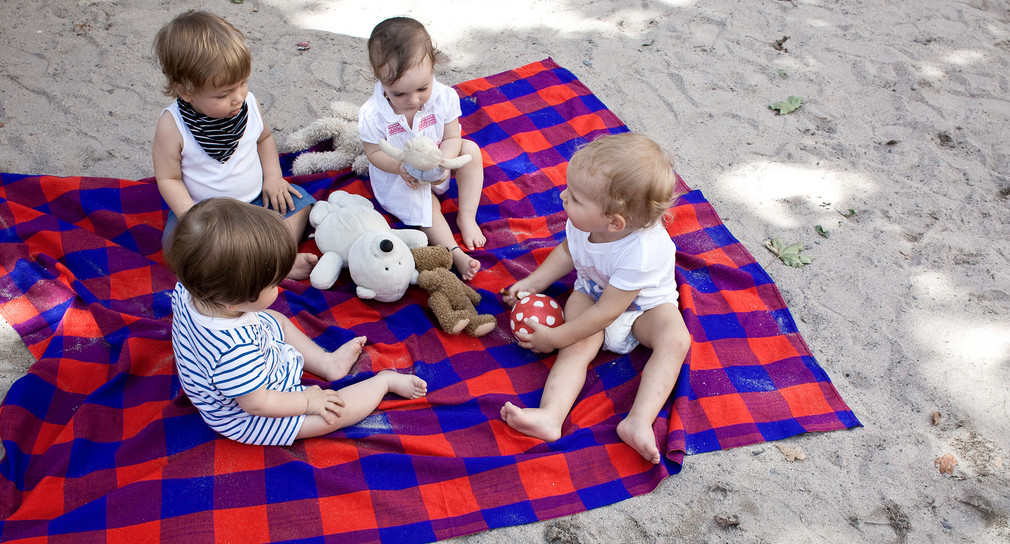 Vier Kleinkinder mit Plüschtieren und einem Ball auf einer Decke