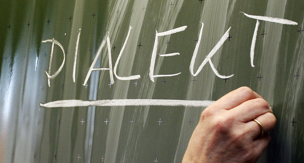 Eine Frau schreibt das Wort „Dialekt“ auf eine Tafel (Bild: © Daniel Karmann/dpa)
