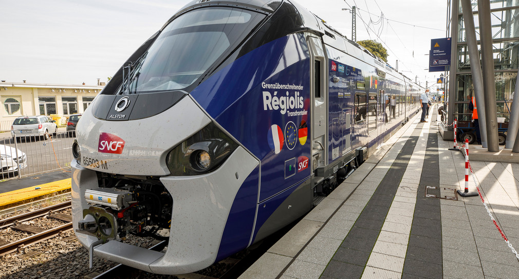 Ein blau-weißes Triebfahrzeug mit der Flagge von Frankreich und Deutschland steht an einem Bahnsteig.