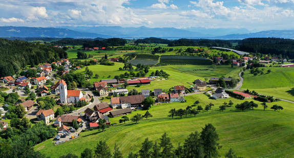 Das beschauliche Dorf Hiltensweiler, ein Teilort von Tettnang, wird von der Abendsonne angestrahlt. Im Hintergrund sind der Bodensee und die Alpen zu sehen. 