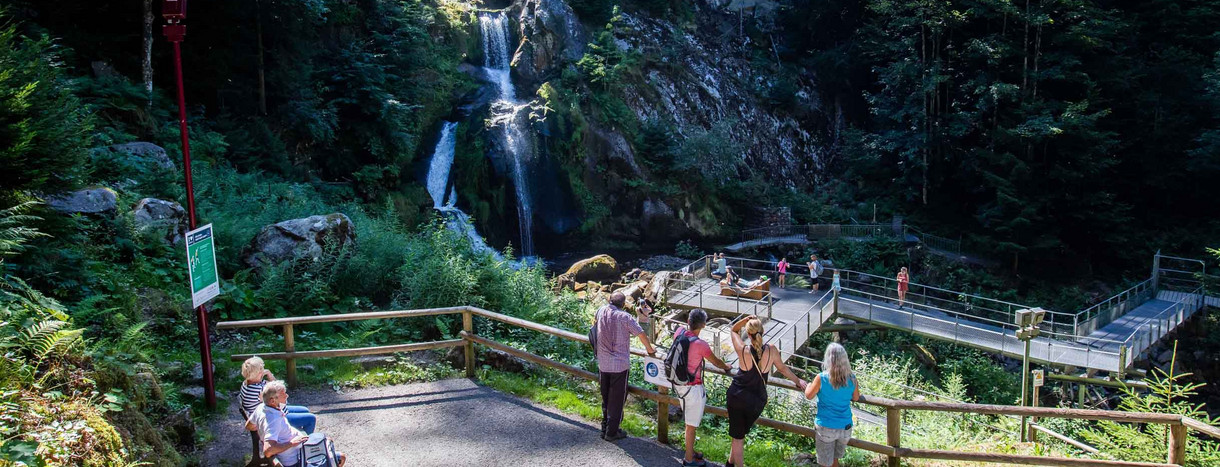 Touristen stehen an einem Geländer und auf einer Aussichtsplattform und blicken auf eine Stufe der Triberger Wasserfälle, über die Wasser fließt.