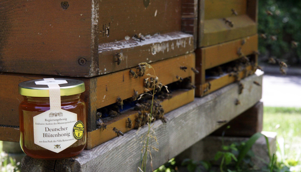 Ein Glas vom Regierungshonig steht vor einem Bienenhaus.