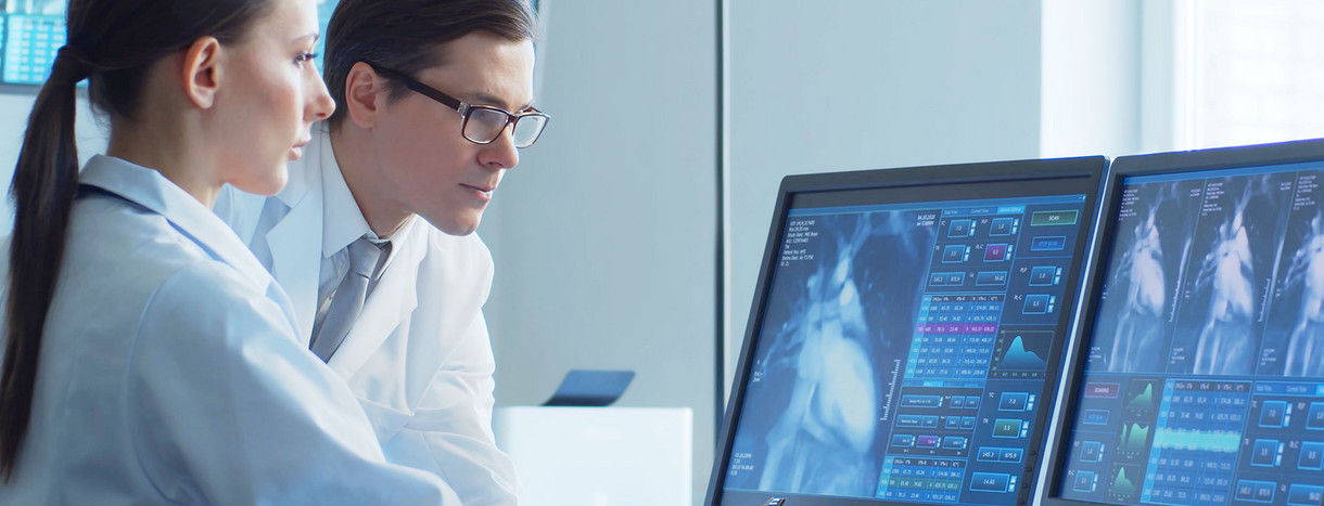 Zwei Mediziner*innen stehen vor Bildschirmen mit MRT-Aufnahmen des Herzens.
