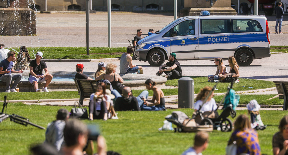 Menschen sitzen auf Wiese mit Polizeiwagen im Hintergrund (Bild: © Christoph Schmidt/dpa)