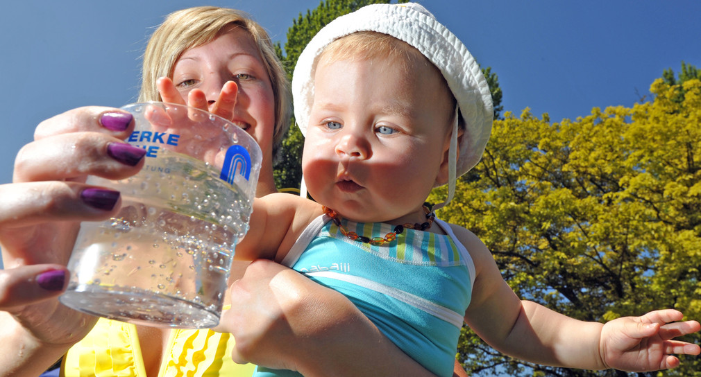 Eine Mutter hält ihr Baby und ein Glas Trinkwasser im Arm (Bild: © dpa).