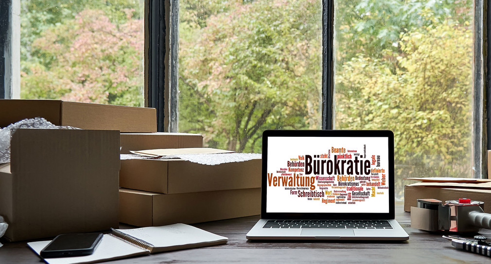 Auf einem Tisch stehen mehrere Pakete und ein Laptop auf dessen Bildschirm eine Tagcloud zum Thema Bürokratie zu sehen ist.