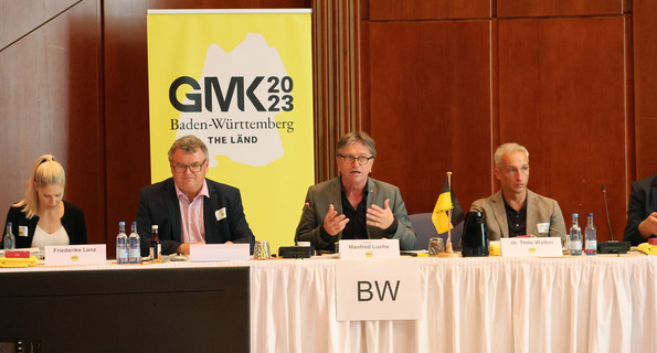 Minister Manne Lucha sitzt mit anderen Teilnehmenden in Konferenzsaal, im Hintergrund eine Stellwand mit GMK-Logo