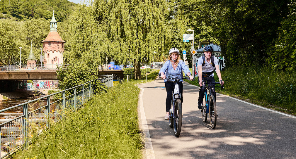 Zwei Radfahrer:innen fahren einen Weg an einem Fluss entlang.