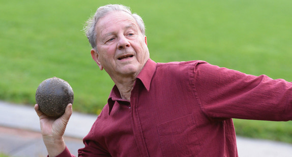 Ein 81-jähriger Mann trainiert auf einem Sportplatz die Disziplin Kugelstoßen. (Foto: © dpa)