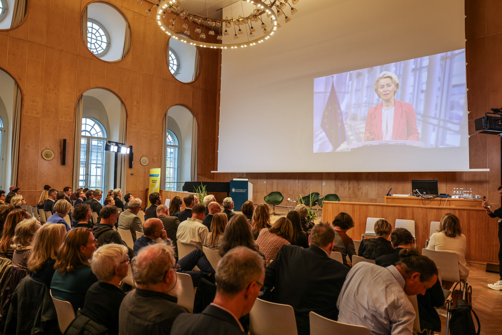 Videobotschaft von Dr. Ursula von der Leyen, Präsidentin der Europäischen Kommission, auf der Leinwand vor den Tagungsteilnehmenden.