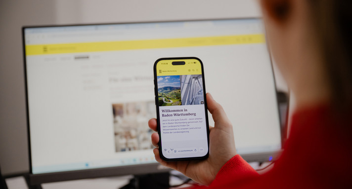 Beschreibung vom Bild: Man sieht eine Hand. Die Hand hält ein Smartphone. Auf dem Smartphone sieht man die Internet Seite des Landesportals Baden-Württemberg.