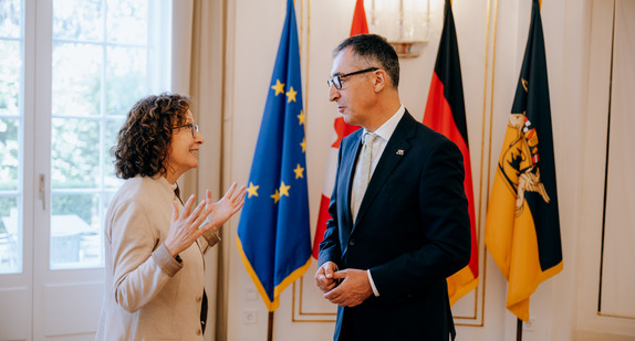 Ministerpräsident Özdemir im Gespräch mit der kanadischen Botschafterin in einem Raum mit großen Fenstern, im Hintergrund die Flaggen der Europäischen Union, Kanadas, Deutschlands und Baden-Württembergs.