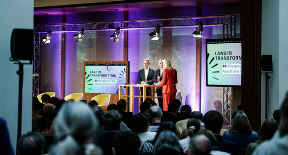 Seitlicher Blick in den Großen Saal über die Köpfe der Gästinnen und Gäste hinweg auf die Bühne, auf der Finanzminister Dr. Danyal Bayaz, Bundesfinanzminister Christian Lindner und die Moderatorin Sonja Álvarez Sobreviela zu sehen sind