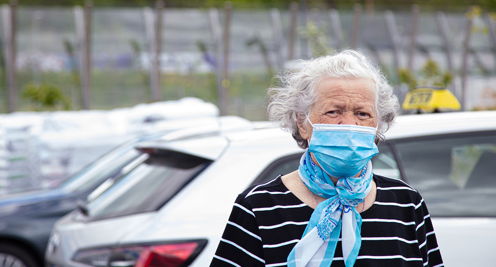 Eine ältere Dame mit einer OP-Maske steht vor einem Taxi auf einem Parkplatz.