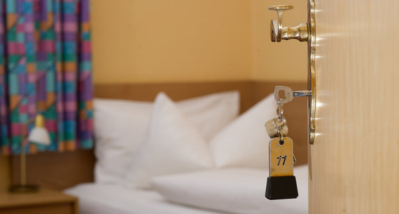 Ein Zimmerschlüssel hängt in einem Gästehaus vor einem Bett im Türschloss. (Bild: © dpa)