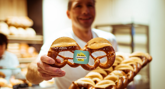 Ein Bäcker hält eine frisch gebackende Zusammenhaltsbrezel in die Kamera