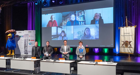 Die Akteurinnen und Akteure der Heimattage Baden-Württemberg 2022 bei der hybriden Auftakt-Pressekonferenz stehen auf einer Bühne beziehungsweise sind auf einer Leinwand zugeschaltet.