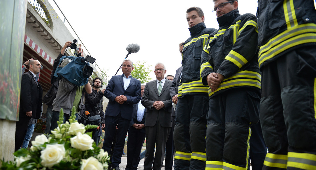 Drei Feuerwehrmänner und Ministerpräsident Winfried Kretschmann (M.) sowie Innenminister Thomas Strobl (l.) stehen in Schwäbisch Gmünd vor der Bahnhofsunterführung am Unglücksort hinter einem Blumenkranz. (Foto: dpa)