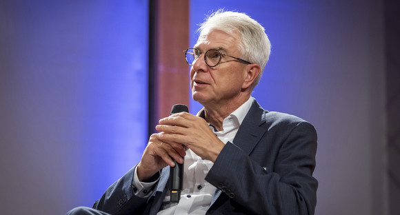 Nahaufnahme von Christian Rauch auf dem Podium. Herr Rauch trägt ein weißes Hemd und ein dunkelblaues Sakko. In den Händen hält er ein Mikrofon. Er schaut, durch seine Brille, nachdenklich nach oben