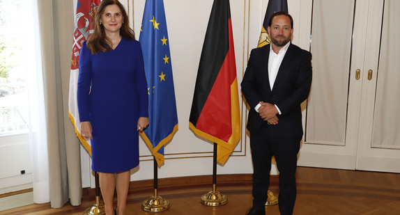 Staatssekretär Florian Hassler (r.) und die serbische Botschafterin Snežana Janković (l.) stehen in der Villa Reitzenstein in Stuttgart vor Fahnen.