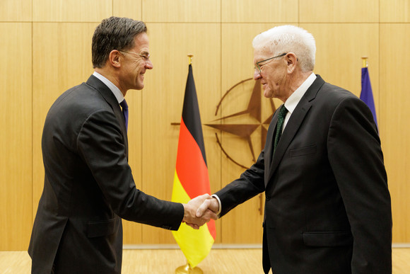 NATO-Generalsekretär Mark Rutte (links) und Ministerpräsident Winfried Kretschmann (rechts) geben sich die Hand. Im Hintergrund stehen Fahnen.
