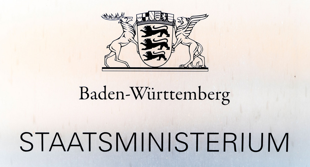 Auf einem silbernen Schild ist in Schwarz das baden-württembergische Landeswappen und die Aufschrift „Baden-Württemberg“ sowie „Staatsministerium“ zu lesen.